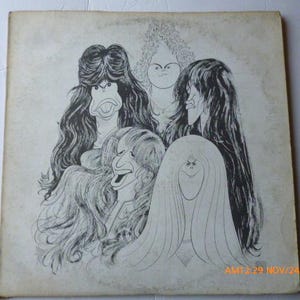 May include: Vintage album cover featuring a black and white cartoon illustration of five stylized faces with exaggerated features and long hair. The artwork has a sketched appearance and is set against a faded, off-white background.