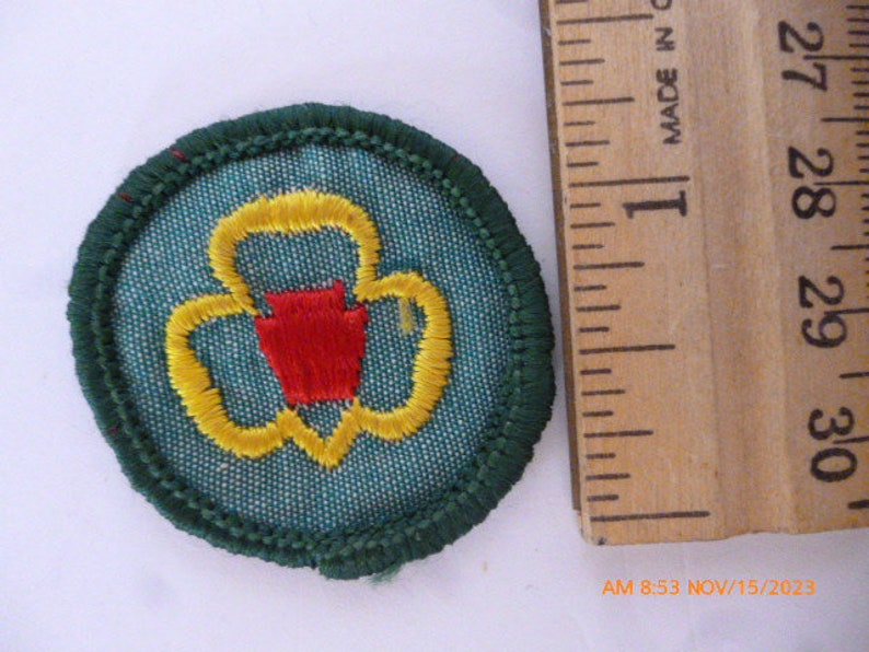 K&ouml;nnte beinhalten: Ein gr&uuml;ner Stoff-Aufn&auml;her mit einer gelben gestickten Blume mit roter Mitte. Die Blume ist eine stilisierte Version des Girl Scout-Symbols.