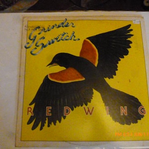May include: Vintage album cover featuring a stylized illustration of a Red-winged Blackbird in flight. The bird is black with orange and red accents, set against a yellow background. The album title is "Grinder Switch".