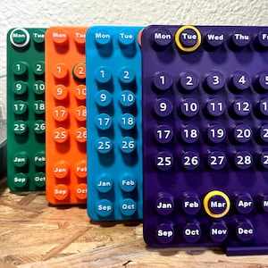 Peut inclure: Gros plan sur quatre calendriers modulaires colorés. Chaque calendrier est d'une couleur différente : vert, orange, bleu et violet. Les calendriers affichent les jours du mois et les mois de l'année. Le mois en cours est mis en évidence en jaune.
