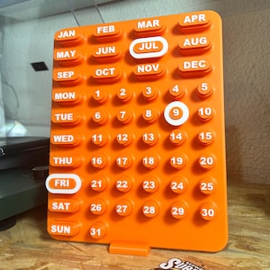 May include: A bright orange perpetual calendar with white lettering. The calendar displays July and the 9th day highlighted in white. The days of the week and months are also labelled in white. The calendar is made of a solid, matte material.