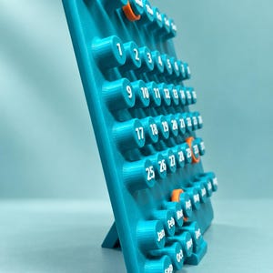 May include: A teal 3D-printed perpetual calendar featuring raised cylindrical buttons with white printed numbers and month abbreviations. The calendar includes select orange-colored buttons for highlighting specific dates and months.