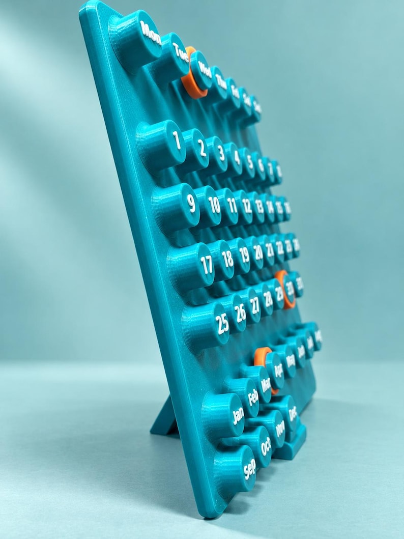 May include: A teal 3D-printed perpetual calendar featuring raised cylindrical buttons with white printed numbers and month abbreviations. The calendar includes select orange-colored buttons for highlighting specific dates and months.