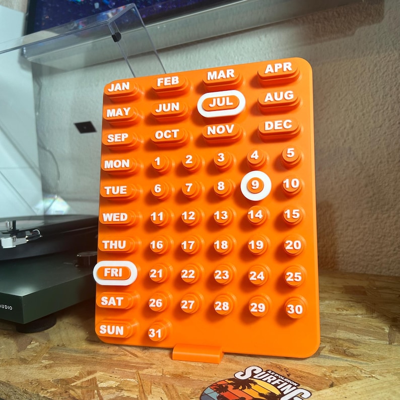 May include: A bright orange perpetual calendar with white lettering. The calendar displays July and the 9th day highlighted in white. The days of the week and months are also labelled in white. The calendar is made of a solid, matte material.
