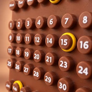 May include: A brown 3D-printed wall calendar featuring raised circular buttons for each day, month, and day of the week. The buttons display white text and numbers. Two yellow rings are placed around the buttons for Friday and the 15th.