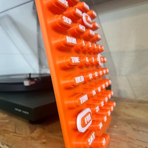 May include: An orange calendar with white lettering and a unique, three-dimensional design. The days of the week and dates are displayed in a clear, easy-to-read format. The calendar is made of a durable material and is designed to stand upright.
