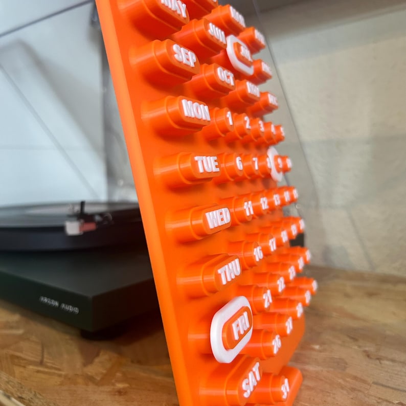 May include: An orange calendar with white lettering and a unique, three-dimensional design. The days of the week and dates are displayed in a clear, easy-to-read format. The calendar is made of a durable material and is designed to stand upright.