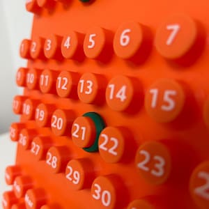 May include: A bright orange perpetual calendar featuring raised circular buttons for each day of the month, numbered 1 through 31 in white text. The top row displays days of the week, and the bottom row shows months, with dark green rings.