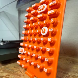 May include: A bright orange perpetual calendar with white text and numbers. The calendar shows months and dates, with the 9th highlighted. The calendar is made of plastic and has a distinctive, three-dimensional design, perfect for a desk.
