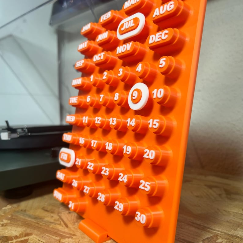 May include: A bright orange perpetual calendar with white text and numbers. The calendar shows months and dates, with the 9th highlighted. The calendar is made of plastic and has a distinctive, three-dimensional design, perfect for a desk.