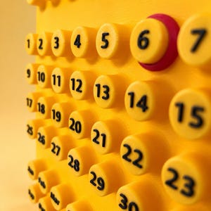 May include: A bright yellow 3D-printed wall calendar featuring raised circular buttons for each day of the month, numbered 1 through 31 in black text. The top row displays days of the week, and the bottom section lists months in black text.