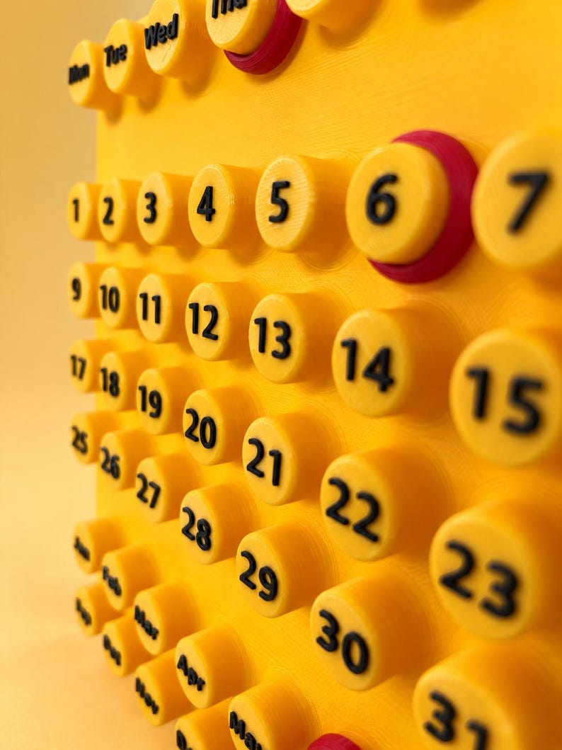 May include: A bright yellow 3D-printed wall calendar featuring raised circular buttons for each day of the month, numbered 1 through 31 in black text. The top row displays days of the week, and the bottom section lists months in black text.