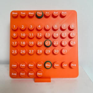 May include: A bright orange square perpetual calendar featuring raised circular buttons with white text. The grid displays days of the week, numbers 1 through 31, and months from January to December. Dark green rings highlight Thu, 21, and May.
