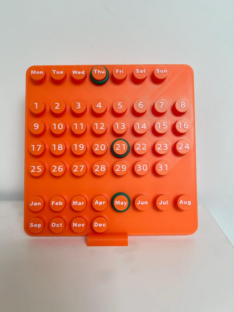 May include: A bright orange square perpetual calendar featuring raised circular buttons with white text. The grid displays days of the week, numbers 1 through 31, and months from January to December. Dark green rings highlight Thu, 21, and May.