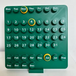 May include: A square green wall calendar featuring raised circular buttons with white text for days of the week, numbers one through thirty-one, and months from January to December. Three buttons are highlighted with yellow rings.
