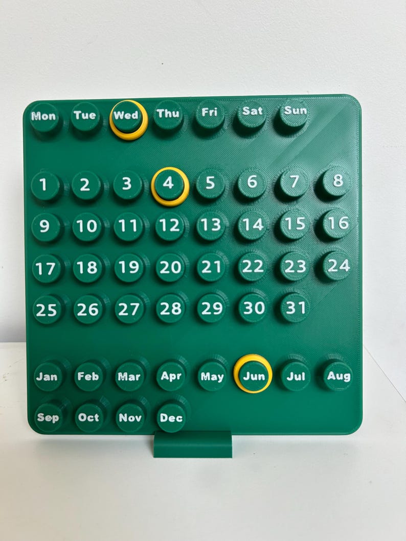 May include: A square green wall calendar featuring raised circular buttons with white text for days of the week, numbers one through thirty-one, and months from January to December. Three buttons are highlighted with yellow rings.
