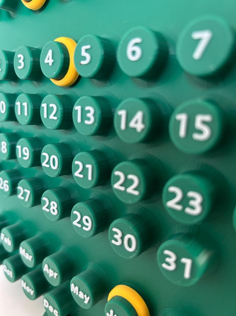 May include: A green wall calendar featuring raised circular buttons for each day of the month, numbered 3 through 31 in white text. A yellow ring highlights the number 4. Below the days, month abbreviations including Feb, Mar, Apr, May, and Dec are visible.