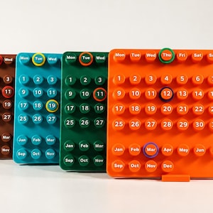 May include: Four rectangular perpetual calendars in brown, teal, green, and orange. Each features raised circular buttons with printed days of the week, numbers 1 through 31, and months from January to December, with select colored rings.