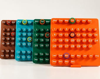 Peut inclure: Quatre calendriers perpétuels rectangulaires de couleur marron, bleu sarcelle, vert et orange. Chacun comporte des boutons circulaires en relief avec les jours, les chiffres de 1 à 31 et les mois, agrémentés d'anneaux colorés.