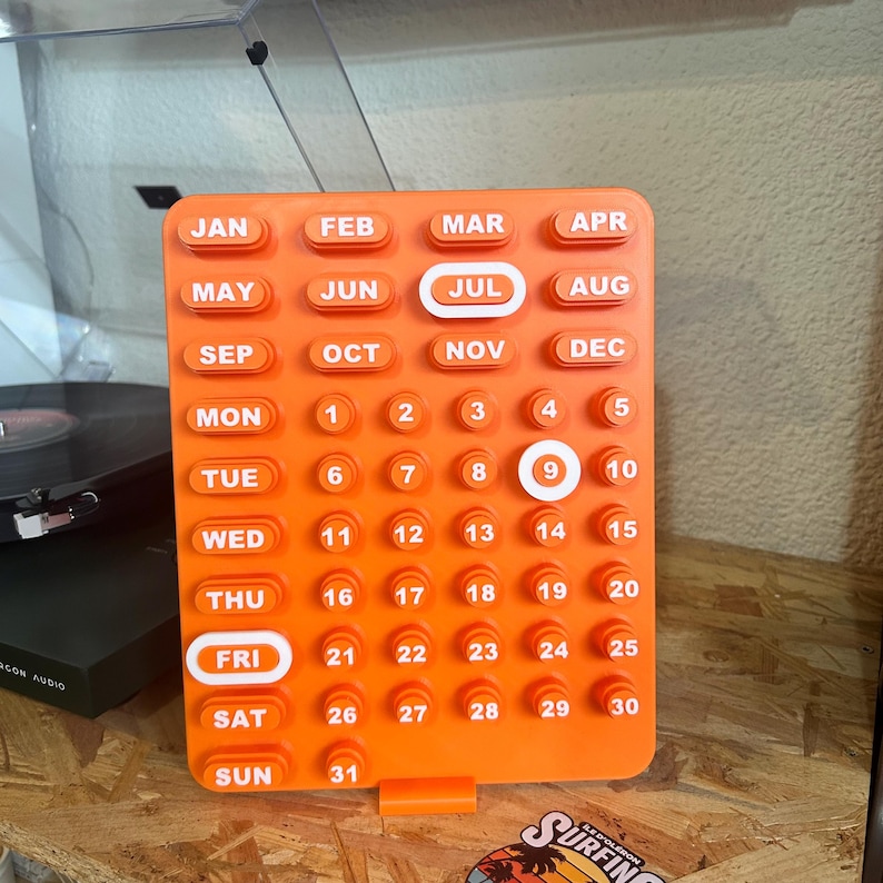 May include: An orange, rectangular perpetual calendar with white text and numbers. The months and days of the week are labeled. The current date is highlighted on the calendar. The calendar is on a wooden surface.