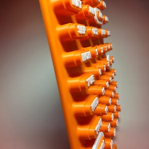 May include: An orange plastic calendar with white lettering. The calendar displays the days of the week and numbers. The calendar is angled, showing the side view of the raised buttons with the dates and days.
