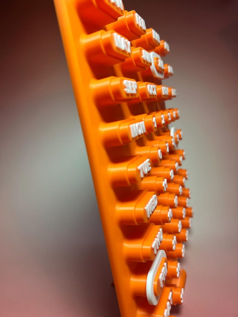 May include: An orange plastic calendar with white lettering. The calendar displays the days of the week and numbers. The calendar is angled, showing the side view of the raised buttons with the dates and days.