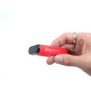 Può includere: Un cinturino rosso per smartwatch con schermo nero è tenuto in mano. Il cinturino riporta la scritta "Smartwatch Market". Il cinturino è realizzato in materiale flessibile e presenta diversi fori per la regolazione.