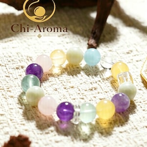 Könnte beinhalten: Ein Perlenarmband mit glatten, runden, mehrfarbigen Edelsteinen in den Farben Violett, Rosa, Gelb, Hellgrün und Blau, durchsetzt mit klaren, transparenten Zwischenperlen. Das Logo Chi-Aroma ist in der oberen linken Ecke zu sehen.