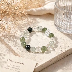 May include: A beaded bracelet featuring a mix of translucent clear quartz beads, dark green moss-like inclusions, solid dark green beads, and a single clear square cube bead. The bracelet is arranged in a circular shape on a textured stone surface.