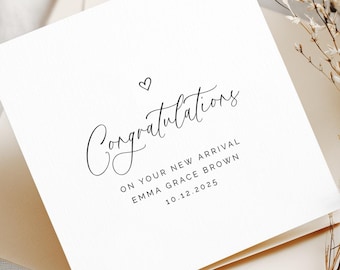May include: A white square card with the word "Congratulations" in elegant script, a small heart, and the text "ON YOUR NEW ARRIVAL EMMA GRACE BROWN 10.12.2025". The card rests on a cream-colored envelope, with dried flowers in the background.