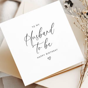May include: A white square greeting card with the text "TO MY Husband To be HAPPY BIRTHDAY" in black script. The card is on top of a cream-colored envelope, with dried flowers in the background.