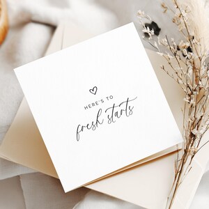 May include: A square white greeting card featuring a small black heart icon above the text "HERE'S TO fresh starts" in a combination of sans-serif and script fonts. The card is placed on top of a matching envelope, accompanied by dried beige grass.