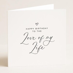 May include: A white, square greeting card with the message "HAPPY BIRTHDAY TO THE Love of my Life" in black script. A small heart is above the text. The card is folded.