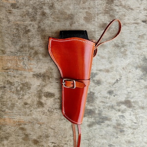 May include: A handcrafted, reddish-brown leather holster with white stitching. The holster features a belt loop with a buckle and a strap. The top of the holster is black. The design is for a handgun.