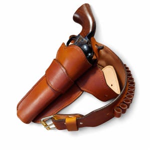 Peut inclure: Un étui en cuir marron contenant un revolver avec un canon en métal sombre et une poignée en bois poli. L'étui est fixé à une ceinture en cuir assortie dotée d'une série de boucles intégrées pour les munitions et d'une boucle en métal argenté.