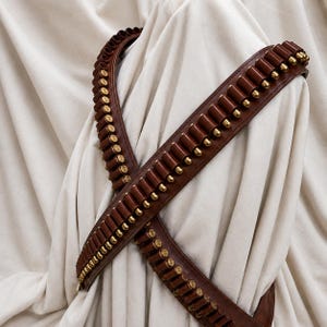 Könnte beinhalten: Ein brauner Leder-Bandolier mit mehreren genähten Schlaufen, die messingfarbene Metallpatronen halten. Der Riemen ist in einer gekreuzten Konfiguration angeordnet und zeigt das sich wiederholende Muster der Munitionshalter entlang der gesamten Länge.