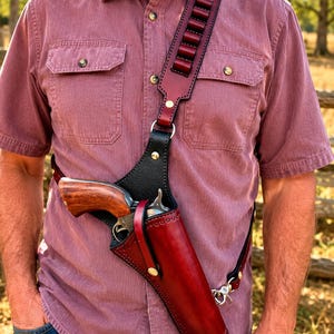 May include: A leather shoulder holster featuring a deep red finish with black leather accents and brass hardware. The strap includes six integrated loops for ammunition, and the holster secures a revolver with a polished wooden grip.