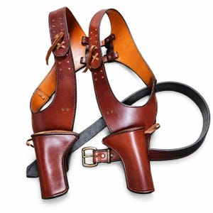 May include: A brown leather double shoulder holster rig featuring two molded holsters, adjustable straps with lacing details, and a black leather belt with a brass-toned metal buckle. The construction includes visible stitching and metal rivets.