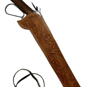 May include: A brown leather rifle scabbard with intricate floral tooling. The scabbard has a dark wooden handle and a metal loop. A black leather strap is attached. The overall design suggests a Western or cowboy theme.