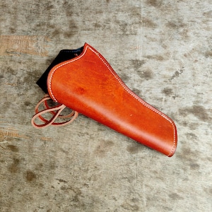 Op de afbeelding: Een handgemaakte, bruin lederen holster met witte stiksels. De holster heeft een gebogen vorm en een lus om te bevestigen. De binnenkant is zwart. De holster is ontworpen om een vuurwapen te bevatten.
