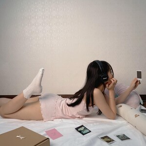 May include: A person wearing a pink top and shorts, lying on a white sheet, wearing black headphones. Several small photos and a brown box with a bow are on the sheet. A pink card is also visible.