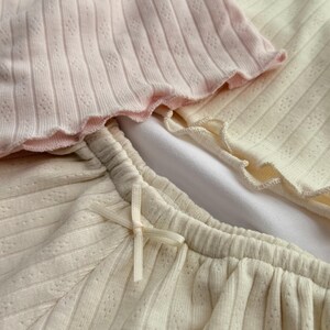 May include: A close-up shot of a set of ribbed knitwear in pastel shades. The set includes a pale pink top with a scalloped edge, and cream-coloured shorts with a delicate bow. The fabric has a subtle textured pattern.