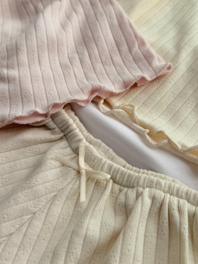 May include: A close-up shot of a set of ribbed knitwear in pastel shades. The set includes a pale pink top with a scalloped edge, and cream-coloured shorts with a delicate bow. The fabric has a subtle textured pattern.