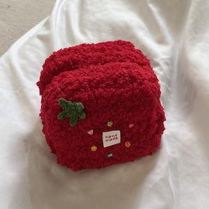 Peut inclure: Un accessoire en forme de fraise rouge en peluche, avec une feuille verte et de petits ornements colorés. Une petite étiquette indique "hand made". L'article est fait d'une matière texturée et floue.