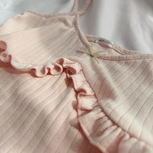 May include: A pale pink camisole top with frilled edges and thin straps. The ribbed fabric has a delicate dotted design. A small label with text is near the neckline. The camisole is made of a soft, lightweight material.