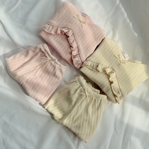 May include: Four sets of baby clothing in pastel shades. The sets include ribbed tops with frilled edges and matching bottoms. Each set is decorated with a small bow. The colours are light pink and cream.