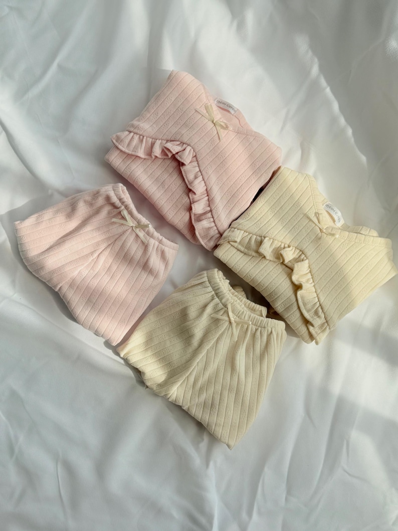 May include: Four sets of baby clothing in pastel shades. The sets include ribbed tops with frilled edges and matching bottoms. Each set is decorated with a small bow. The colours are light pink and cream.