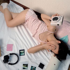 May include: A pale pink pajama set with frilled edges and a white camera. Several polaroid photos, a pair of black headphones, and a pink note are on a white bed. The scene is set against a neutral wall.