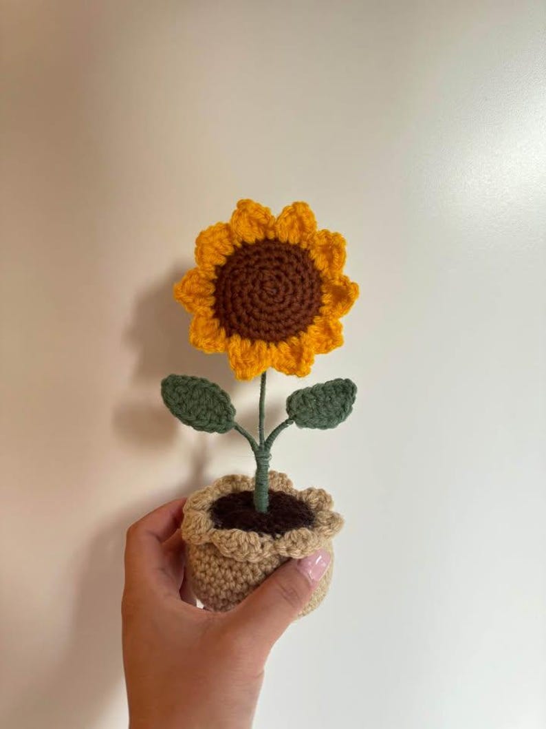 Op de afbeelding: Een handgemaakte gehaakte zonnebloem in een kleine pot. De zonnebloem heeft gele bloemblaadjes en een bruin hart. De groene bladeren en stengel komen uit een beige pot gevuld met bruin materiaal. De pot wordt door een hand vastgehouden.