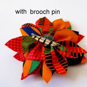 May include: A colorful fabric flower brooch with a safety pin on the back. The flower is made of orange, red, green, black, and blue fabric.